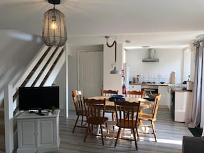 A dining area with a wooden table and chairs, a television on a cabinet, and a kitchen in the background. - Fosfelle Cottages (Bideford)