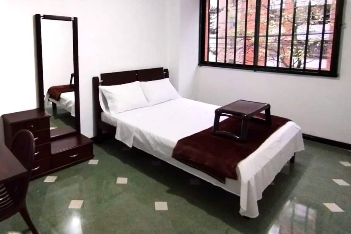 Quarto individual com cama de solteiro, cabeceira de madeira, mesa de cabeceira, guarda-roupa e janela com grades.