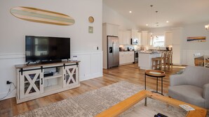 A modern living room with a flat-screen TV, wooden entertainment unit, and a kitchen area with white cabinets and a stainless steel refrigerator. - The Teal Turtle: Beautiful, spacious home with easy beach access (Oak Island)