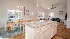 A spacious kitchen with a large island, wooden dining table, and chairs. There are pendant lights, a ceiling fan, and a view of the outside. - The Teal Turtle: Beautiful, spacious home with easy beach access (Oak Island)