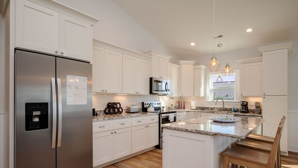 A modern kitchen with white cabinets, a stainless steel refrigerator, a microwave, and a large island with seating. - The Teal Turtle: Beautiful, spacious home with easy beach access (Oak Island)
