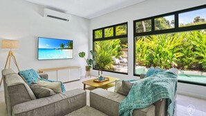 A modern living room with a large window, a ceiling fan, a TV, and a view of a tropical garden. - House Heliconia | Luxury Palm Cove Retreat with Private Pool & Lush Gardens (Palm Cove)