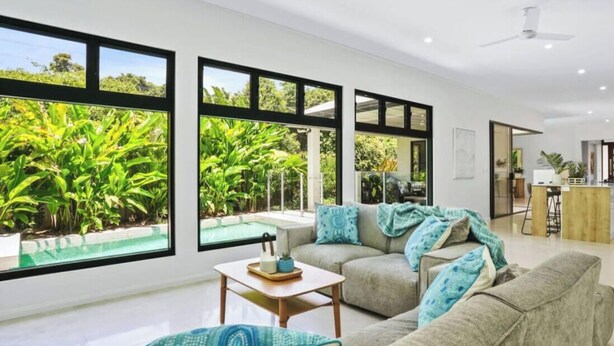 A modern living room with a large window overlooking a pool and lush greenery. - House Heliconia | Luxury Palm Cove Retreat with Private Pool & Lush Gardens (Palm Cove)