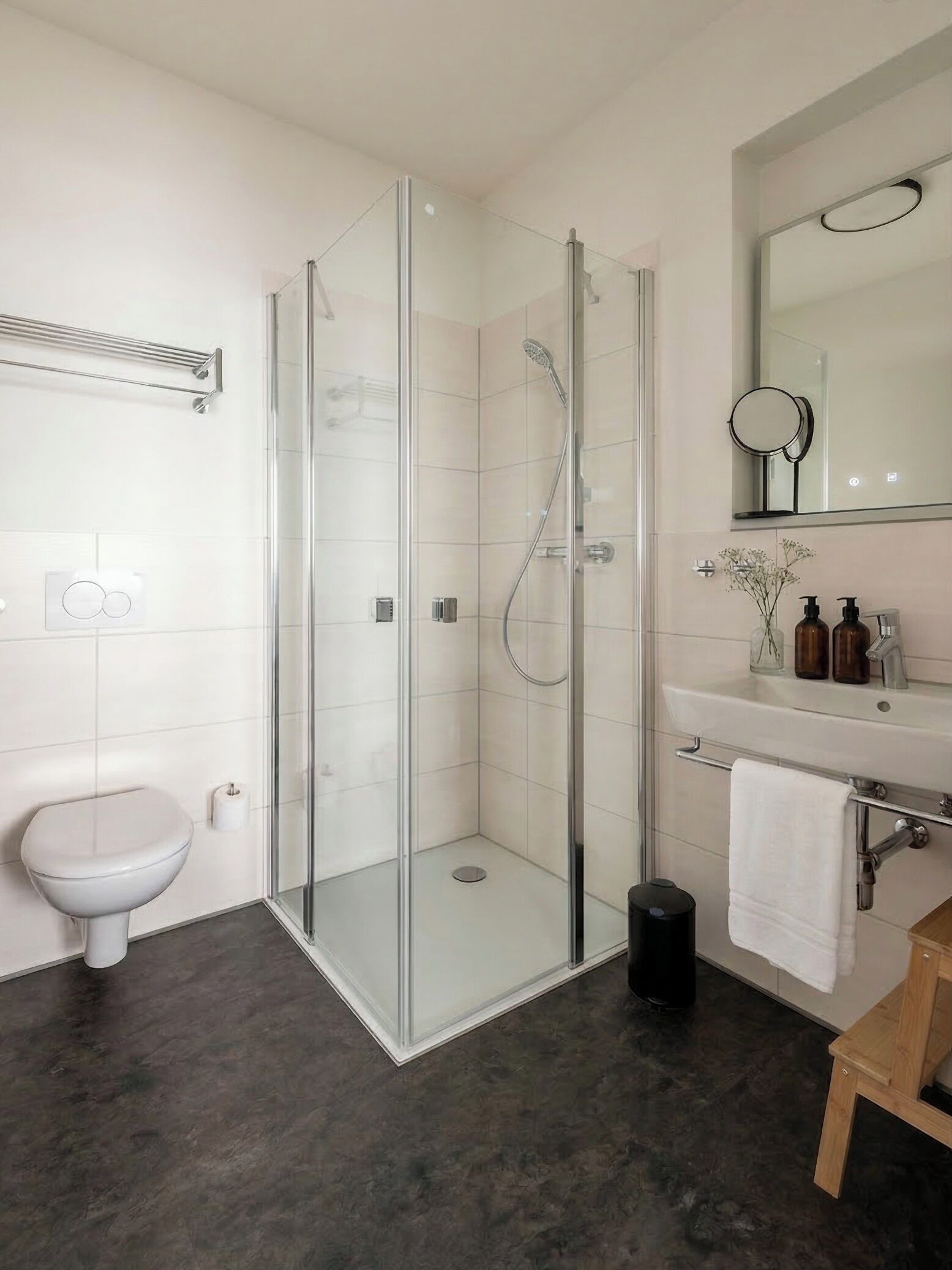 Un baño moderno con ducha envuelta en vidrio, inodoro blanco, lavabo con espejo y un taburete de madera pequeño.