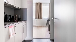 Un pasillo estrecho que conduce a una cocina y comedor con armarios blancos, una tetera negra y una ventana con cortinas.