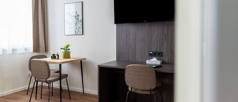 A small, modern room with a desk and two chairs, a flat-screen TV mounted on the wall, and a window with curtains.