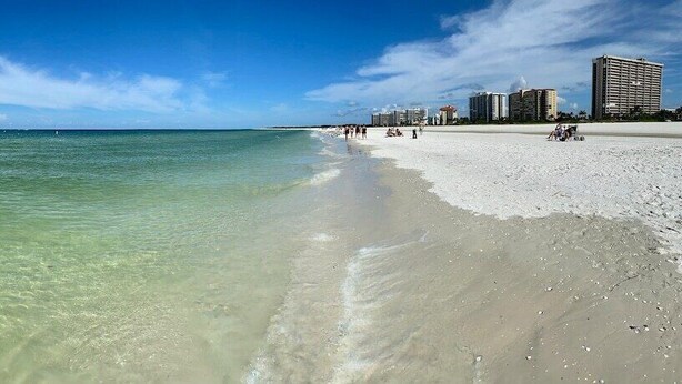 A beach with clear shallow waters, a sandy shore, and buildings in the background.
