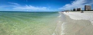 Een strand met helder ondiep water, een zandstrand en gebouwen op de achtergrond.