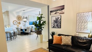 A modern living room with a black leather sofa, a dining area with a white table, and a kitchen visible in the background.