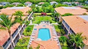 A resort with a central swimming pool, surrounded by villas with red-tiled roofs, palm trees, and a view of the ocean.