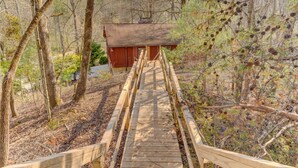 A wooden bridge leading to a red cabin surrounded by trees and pine cones. - Wild Creek Cabin (Mineral Bluff)