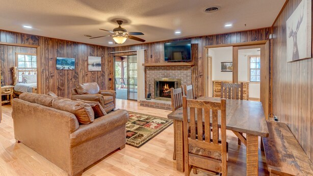 A spacious living room with a fireplace, wooden walls, a ceiling fan, and a TV mounted above it. - Wild Creek Cabin (Mineral Bluff)