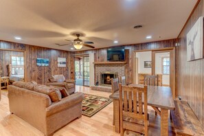A spacious living room with a fireplace, wooden walls, a ceiling fan, and a TV mounted above it.