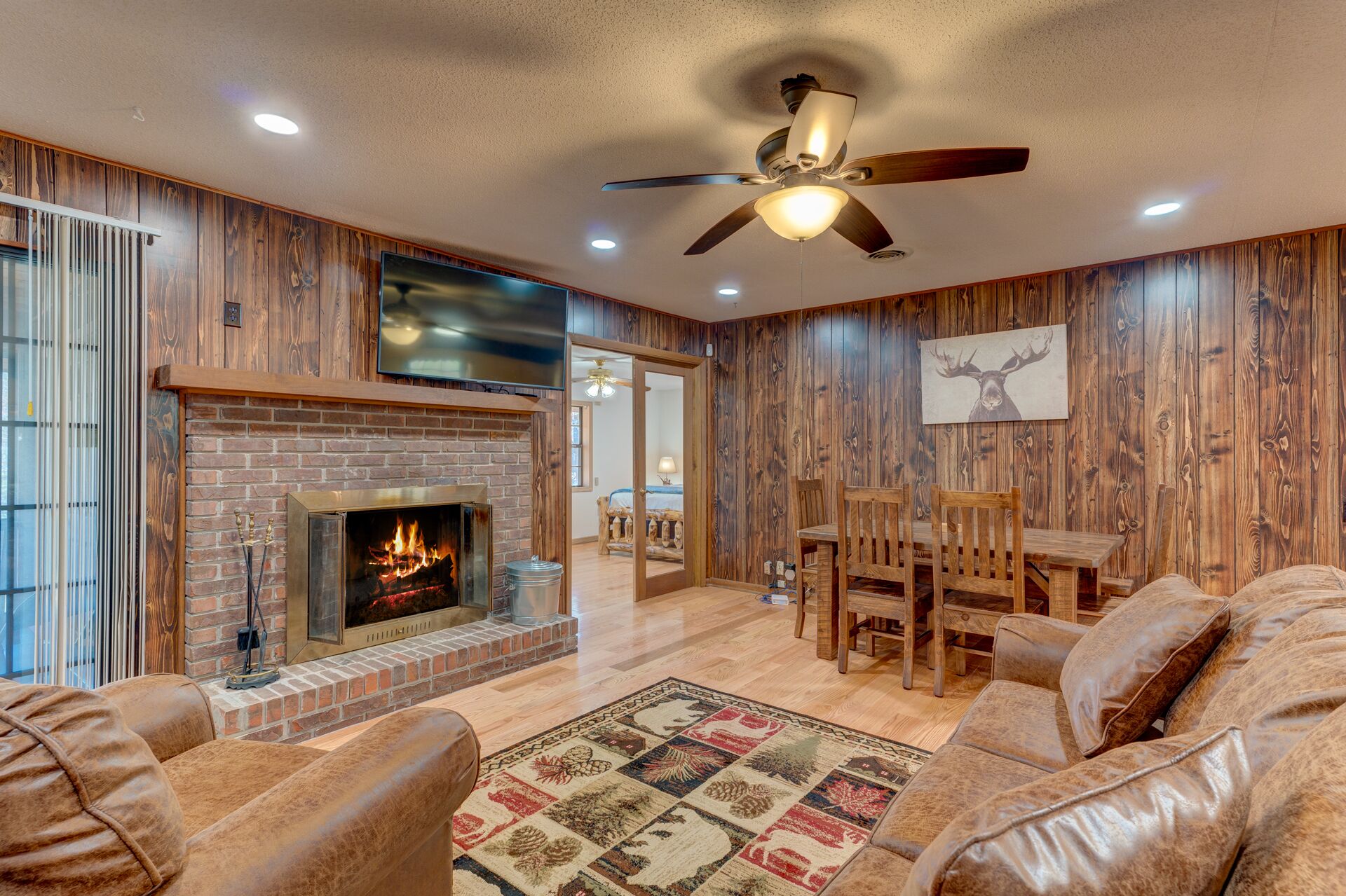 A cozy living room with a brick fireplace, leather sofas, a dining area, and wooden paneling.