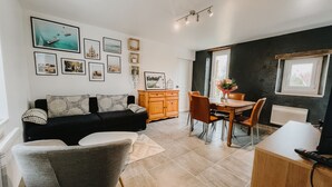 A modern living room with a black sofa, grey armchair, and wooden dining table with chairs. There are framed pictures on the wall and a window with a view. - cottage of the poste (Crouay)