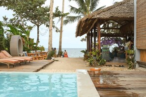 A beachside pool area with lounge chairs, a thatched-roof structure, and a view of the sea.