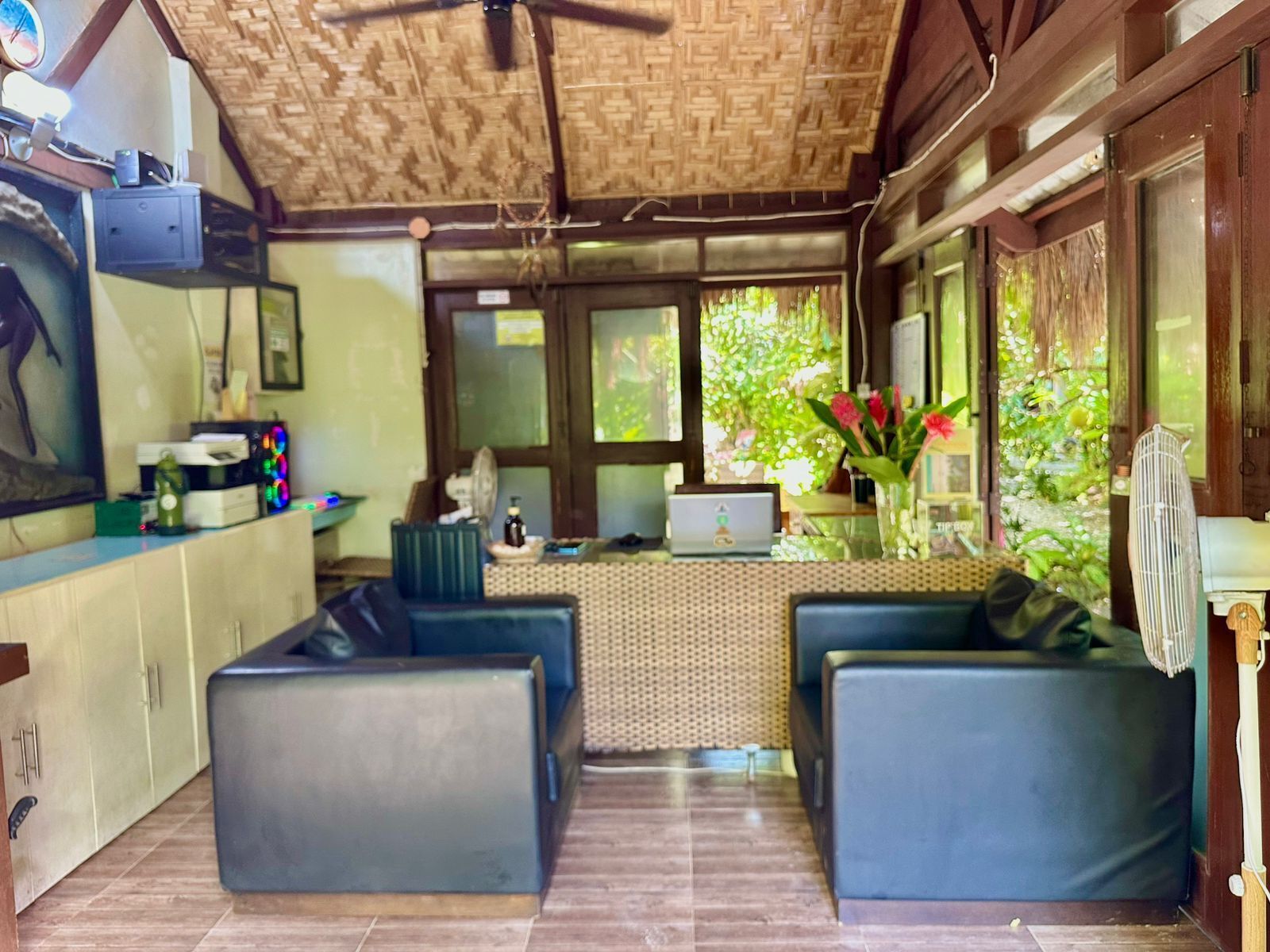 A room with a wooden ceiling, a desk with a computer, two blue sofas, and a view of greenery outside.