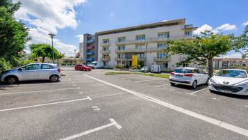 Un parking rempli de voitures, un immeuble à plusieurs étages doté de balcons et un ciel dégagé.