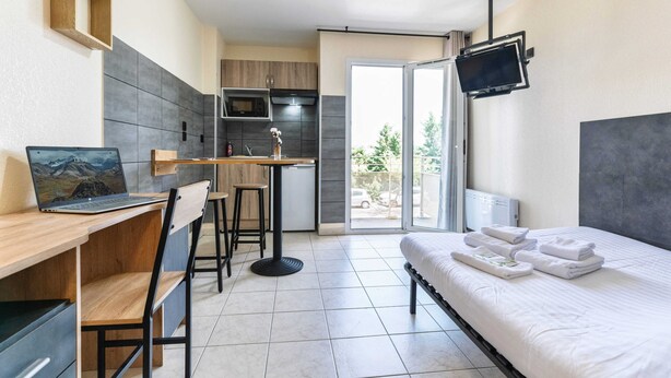 A compact hotel room with a bed, a desk with a laptop, a small kitchen area, and a TV mounted on the wall. - Résidence Columba  (Agen)
