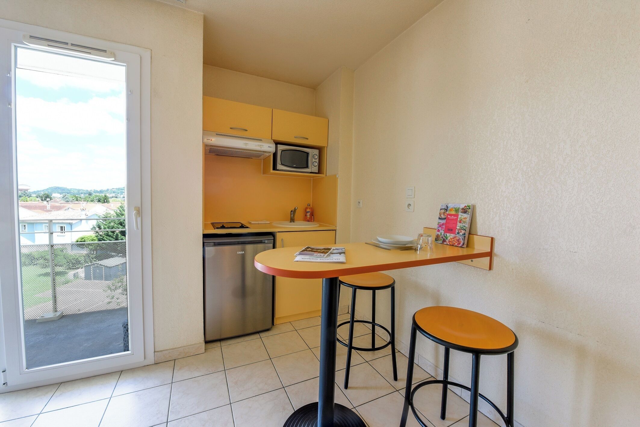 Une cuisine compacte équipée d’un four à micro-ondes, d’une cuisinière et d’un évier, ainsi qu’une petite table avec deux tabourets et une vue sur l’extérieur grâce à une porte coulissante.