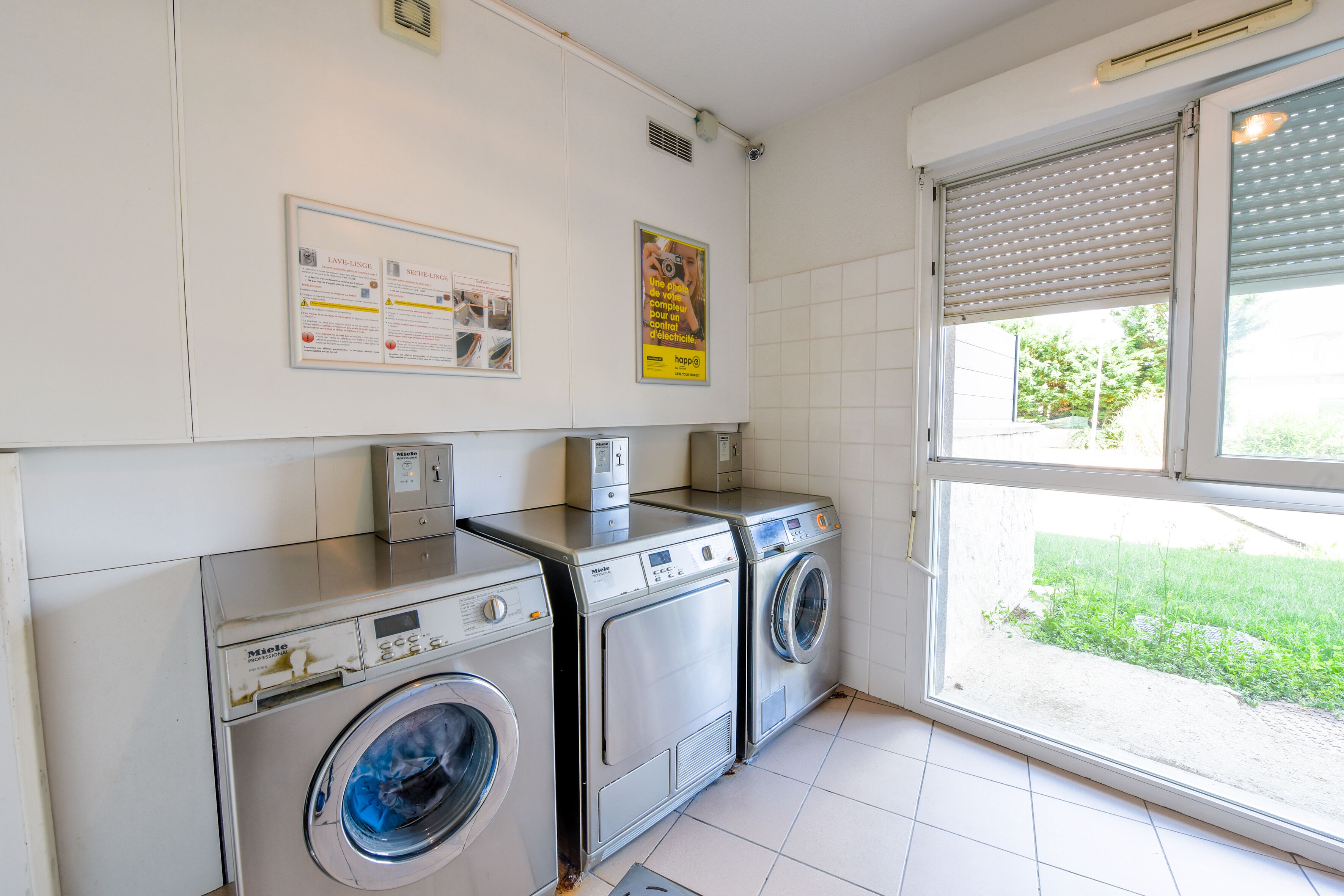 A laundry room with two washing machines and a dryer, a window with blinds, and a wall-mounted information board.