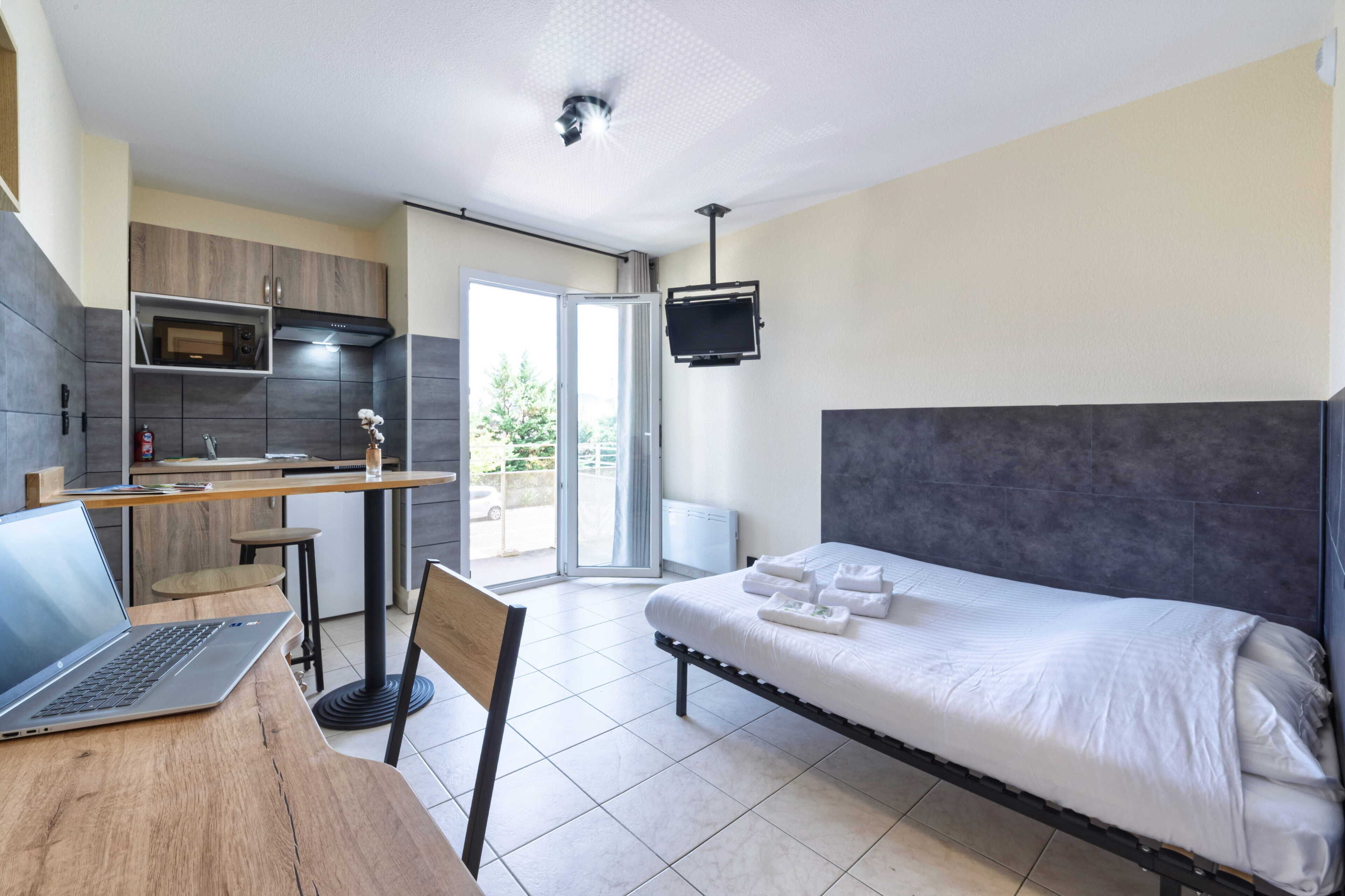 Une chambre d’hôtel moderne équipée d’un lit, d’un bureau avec un ordinateur portable, d’un coin cuisine et d’un téléviseur fixé au mur.