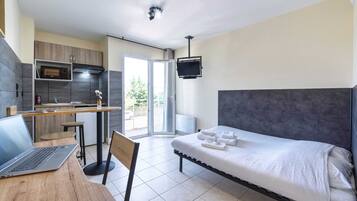 Une chambre d’hôtel moderne équipée d’un lit, d’un bureau avec un ordinateur portable, d’un coin cuisine et d’un téléviseur fixé au mur.