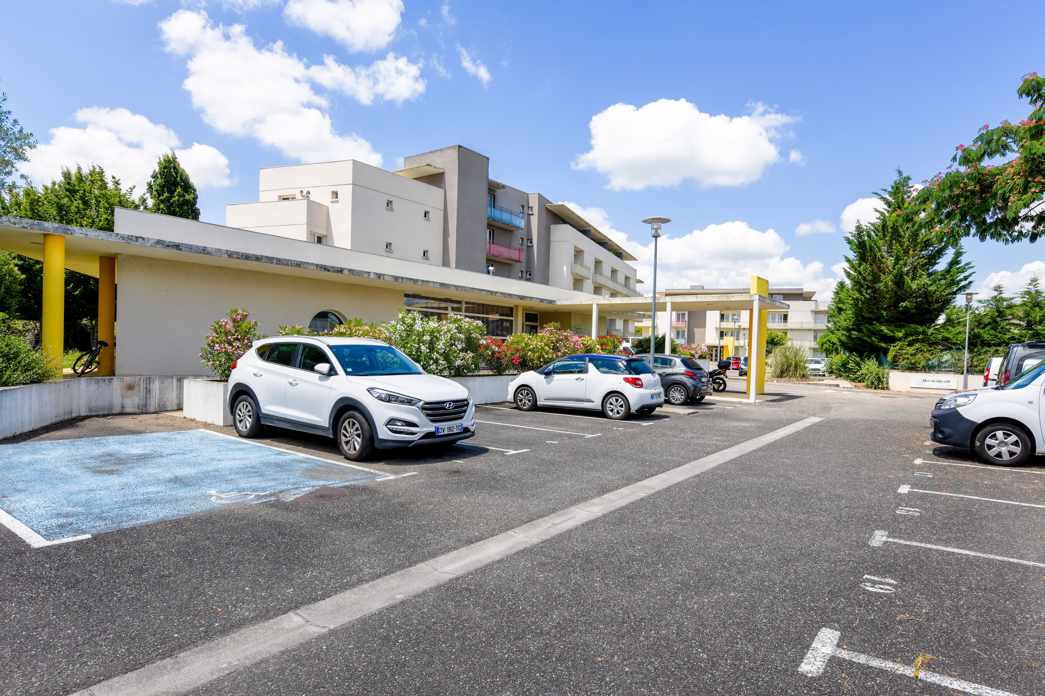 Un parking avec plusieurs voitures, un bâtiment doté d’un toit plat et de balcons, et un ciel bleu dégagé.