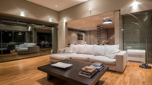 A modern living room with a sectional sofa, a coffee table with books, a floor lamp, and a large window with blinds. - Studio Rio Unique Space With Garden and Garage (Patras)