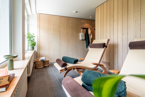 A spa room with wooden panel walls, two lounge chairs with towels, a radiator, a window with a plant, and a shelf with books and a bowl. - stuub hinterzarten (Breitnau)