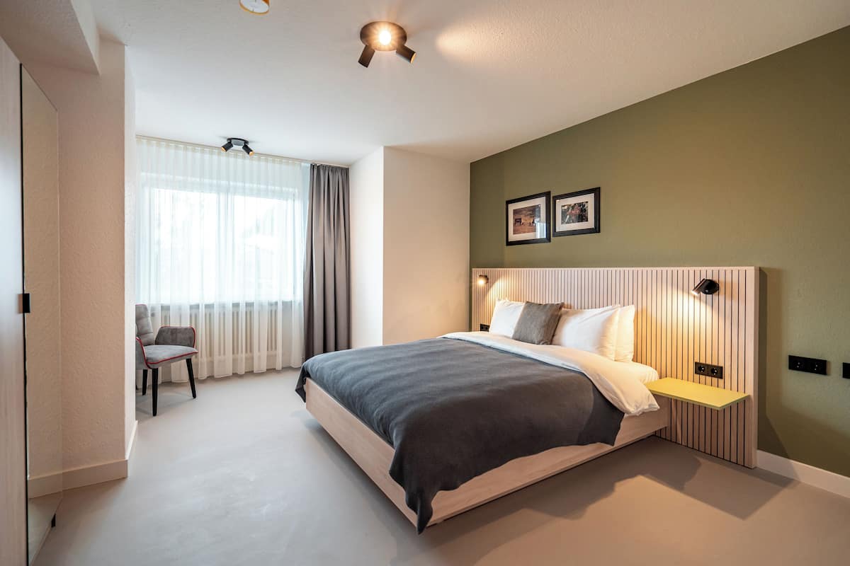 A modern hotel room with a large bed, a bedside table, a chair, a window with curtains, and a wall-mounted light fixture.