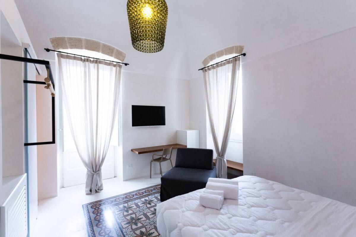 A modern hotel room with a bed, a desk, a chair, a TV, and a patterned rug.