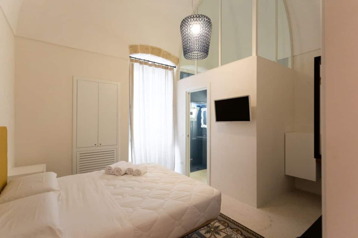 A bedroom with a bed, a wardrobe, a TV, and a bathroom visible through a glass door.