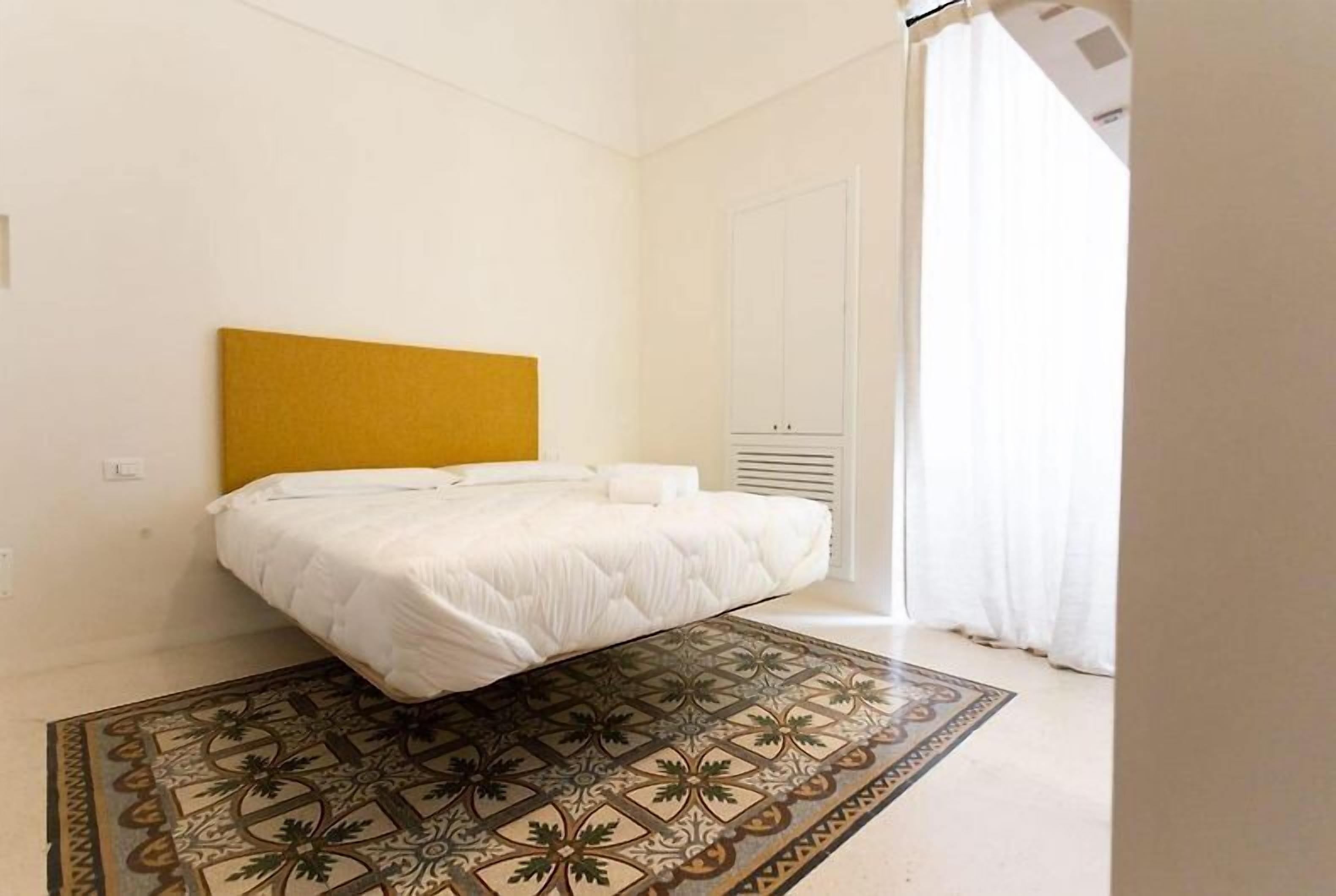 A bedroom with a bed, a headboard, a patterned rug, a white door, and a window with curtains.