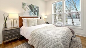 Une chambre à coucher avec un grand lit, deux tables de chevet avec des lampes, un tableau au mur et une fenêtre avec des stores.