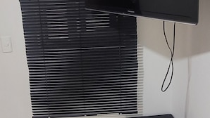 Habitación con televisión de pantalla plana montada, ventana con persianas y un caja de almacenamiento negra.
