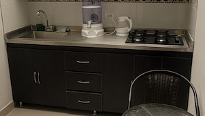 Una cocina compacta con estufa a gas, cafetera y pileta. Se observa una silla y una mesita en primer plano.