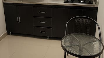 Una cocina compacta con estufa a gas, cafetera y pileta. Se observa una silla y una mesita en primer plano.