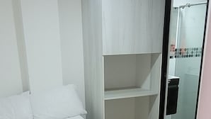 Un dormitorio con cama, una manta amarilla, almohada blanca, cabecera blanca, armario blanco, cabina de ducha de vidrio y piso con baldosas.