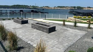 Un patio frente al agua con asientos de piedra, barandilla de metal y vistas al agua y edificios distantes.