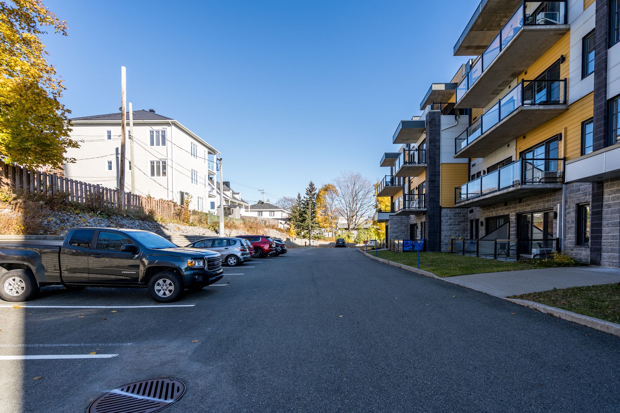 Straßenansicht eines Wohngebiets mit modernen Mehrfamilienhäusern, parkenden Autos und blauem Himmel.