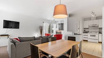 A modern living room with a grey sofa, a wooden dining table, and black chairs. There is a TV mounted on the wall and a kitchen area visible in the background.