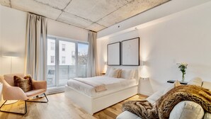 A modern bedroom with a large bed, a sofa, and a chair. There are two framed pictures on the wall and a window with curtains.