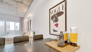 A modern hotel room with a large window, a bed, a dining table with a coffee machine, and a framed magazine on the wall.
