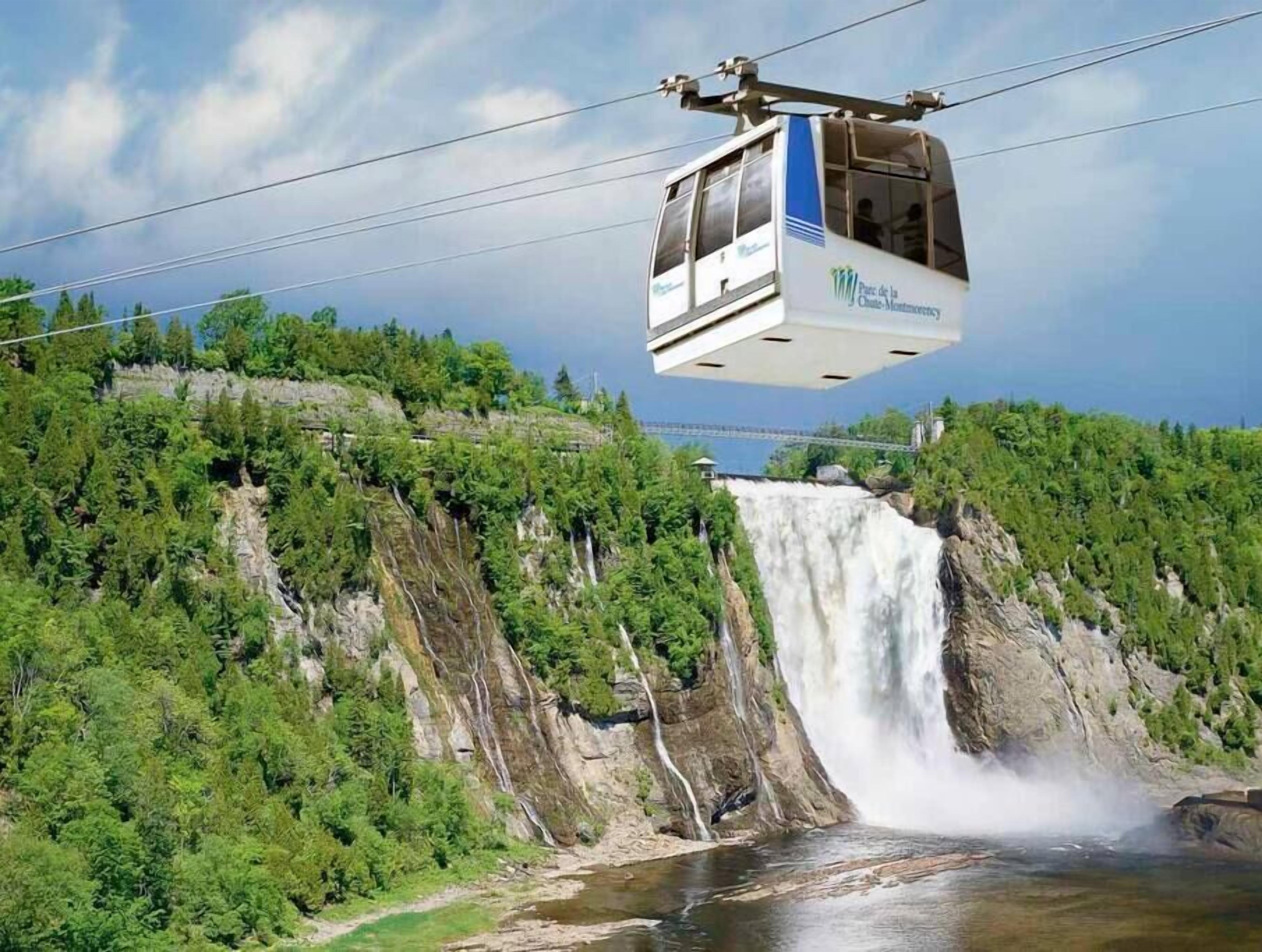 En linbana med en logotyp, som färdas över ett vattenfall med en flod nedanför och en skogbeklädd sluttning.