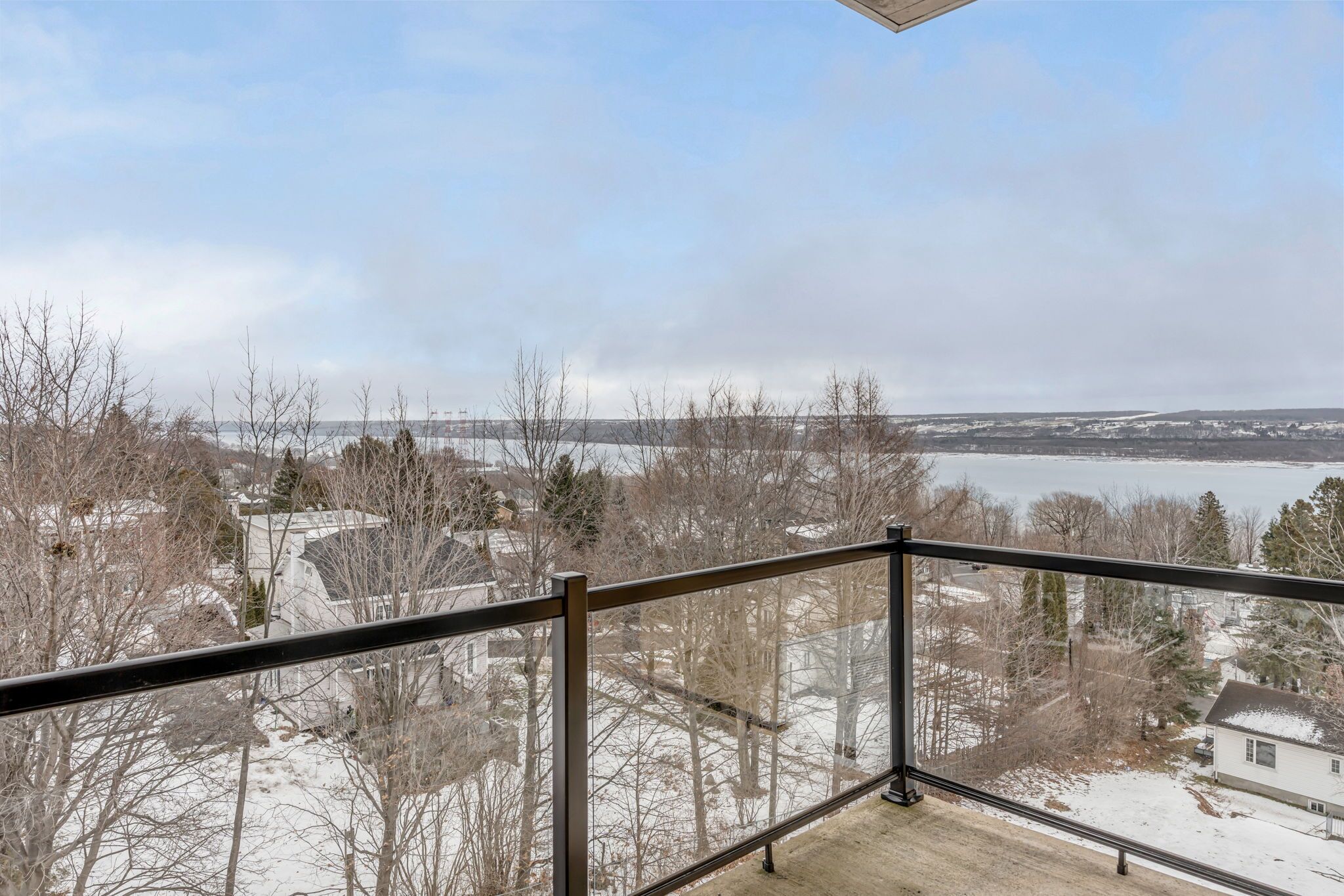 Ein Balkon mit Metallgeländer, der auf eine verschneite Landschaft mit Bäumen und einem Gewässer blickt.