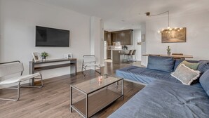 Een moderne woonkamer met een bank, een glazen salontafel, een televisie en een keuken op de achtergrond.