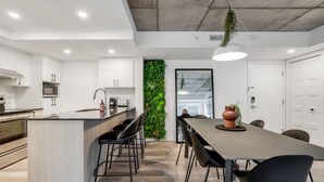 A modern kitchen with white cabinetry, a central island with bar stools, a hanging plant, and a dining area with a long table and chairs.