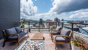 A rooftop terrace with a sofa, chairs, and a table, overlooking a cityscape with buildings and a bridge.