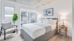 A modern bedroom with a large bed, a chair, a bedside table, and a potted plant.